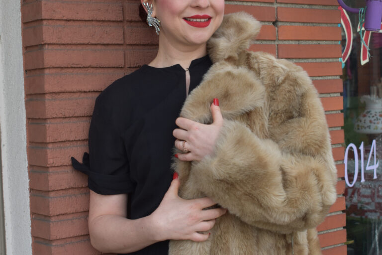 a woman in a black dress holds a fur coat while leaning on a brick wall