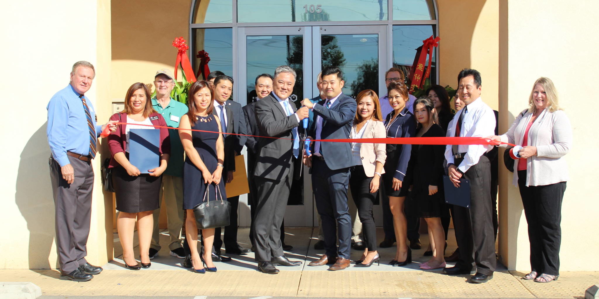 a group of people holding a red ribbon while two men in the center hold a pair of giant scissors