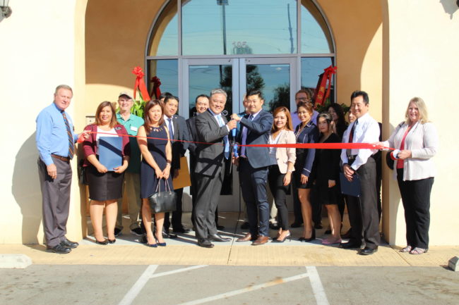 a group of people holding a red ribbon while two men in the center hold a pair of giant scissors