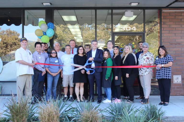 a group gathered in front of a building and a man in the center holds a paid of giants scissors while others hold a red ribbon