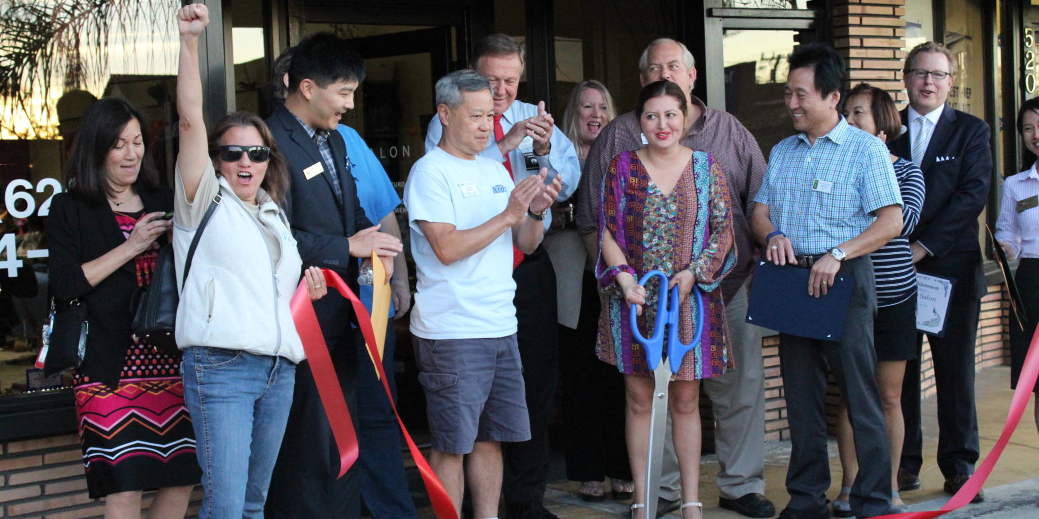 a group of people stands beside a woman holding a large pair of scissors and there is a cut ribbon on the ground