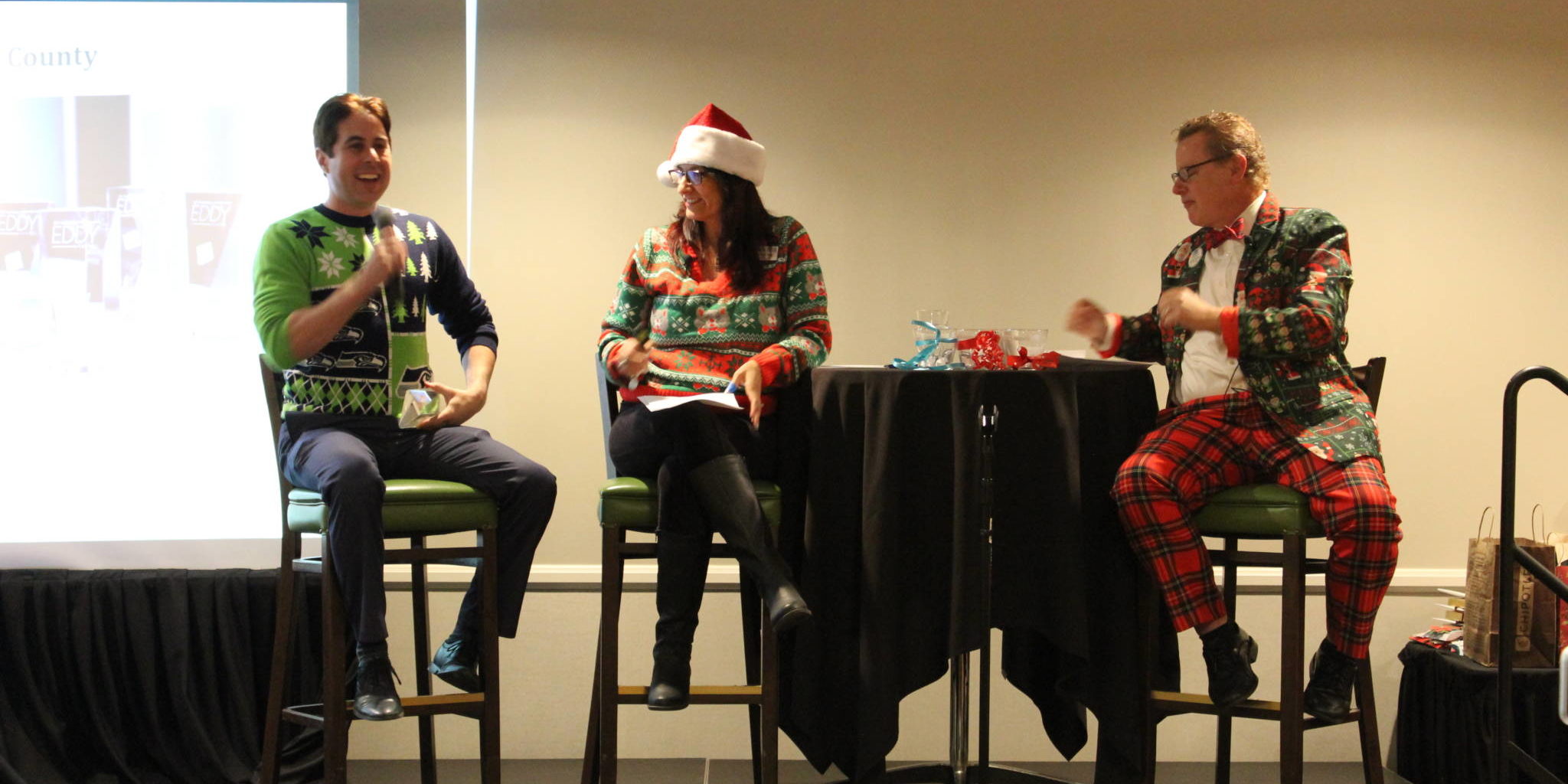 a woman in a santa hat sits between two men dressed in funny holiday attire on a stage