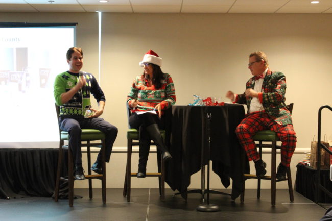 IMG_6455 a woman in a santa hat sits between two men dressed in funny holiday attire on a stage
