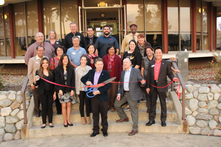 a group of people standing on the back steps of the Arcadia Chamber building and one is holding giant scissors