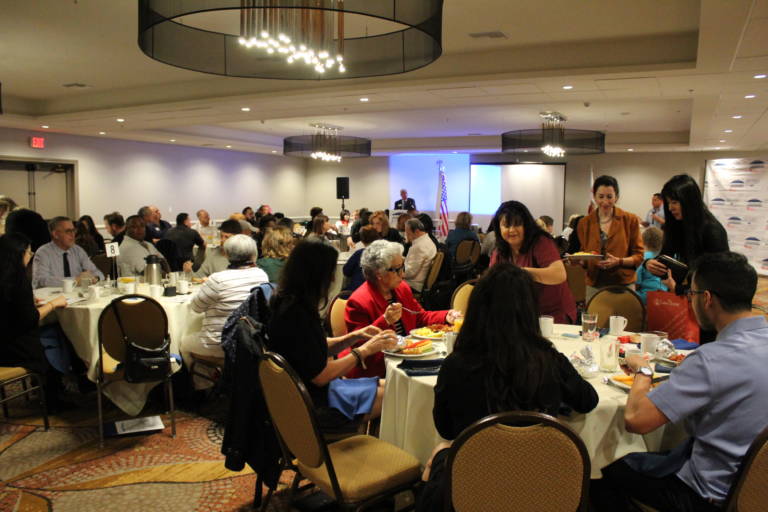 IMG_8778 a room full of people seated and standing around round tables with a projector screen in the background