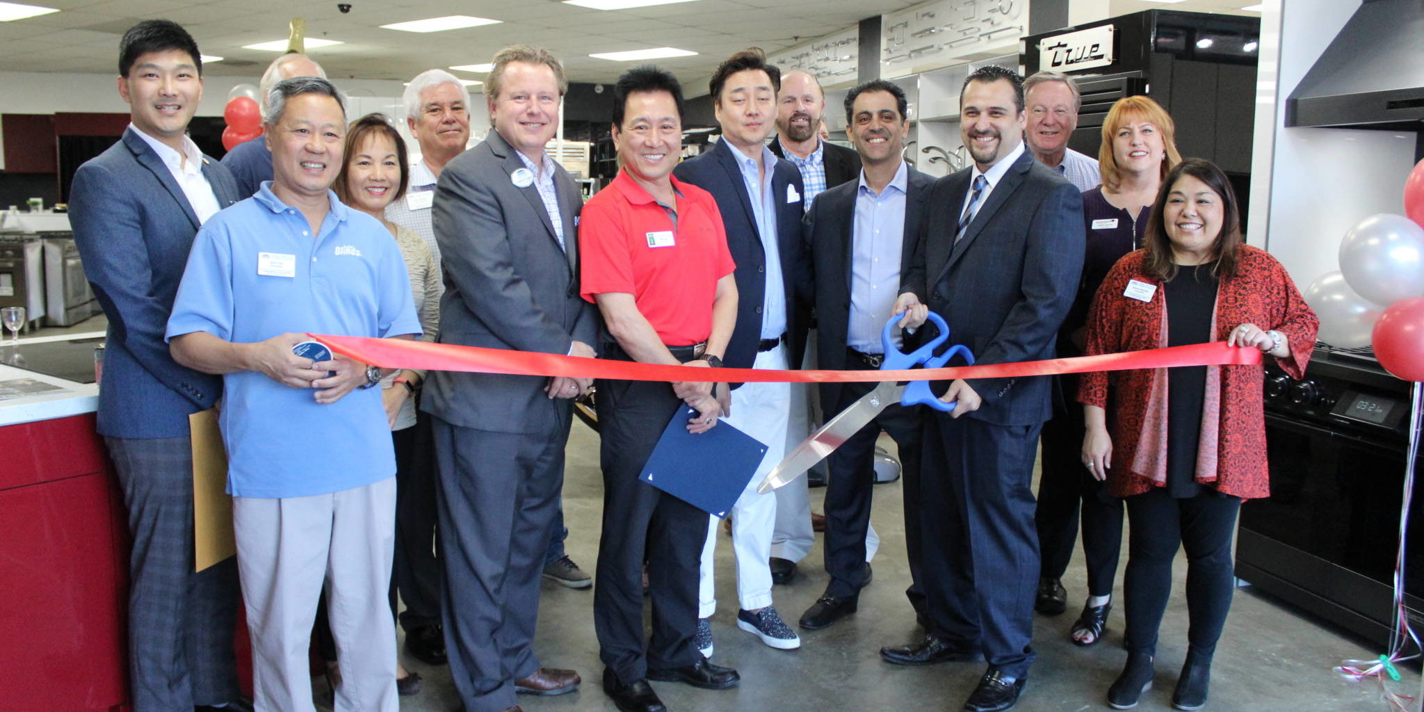 a group of woman and men holding a ribbon and one holds giant scissors as they get ready for a ribbon cutting