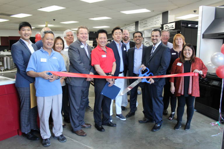 a group of woman and men holding a ribbon and one holds giant scissors as they get ready for a ribbon cutting