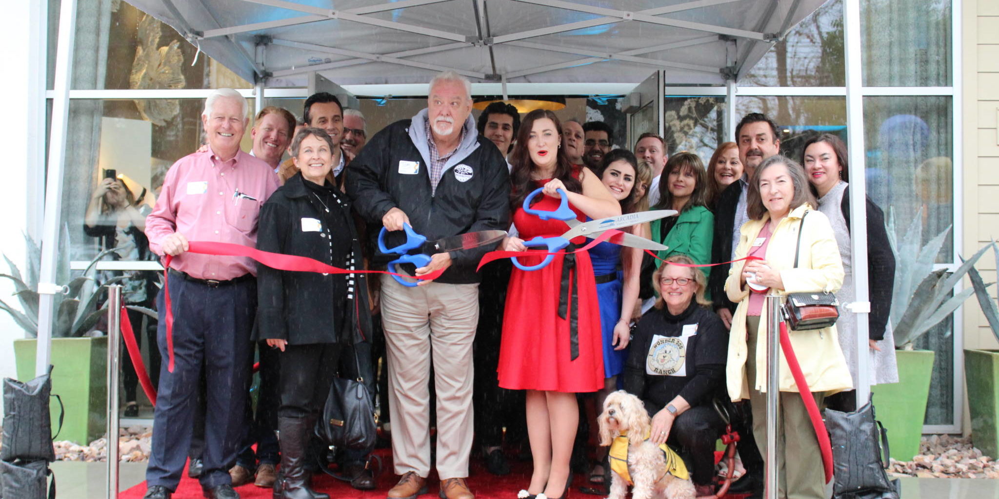 IMG_3390 a bunch of people gathered tightly under an awning cutting a ribbon with a dog