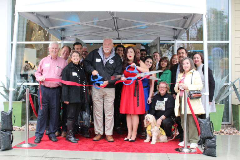 a bunch of people gathered tightly under an awning cutting a ribbon with a dog