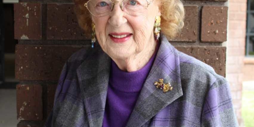 an older woman in a purple plaid coat leaning against a brick wall