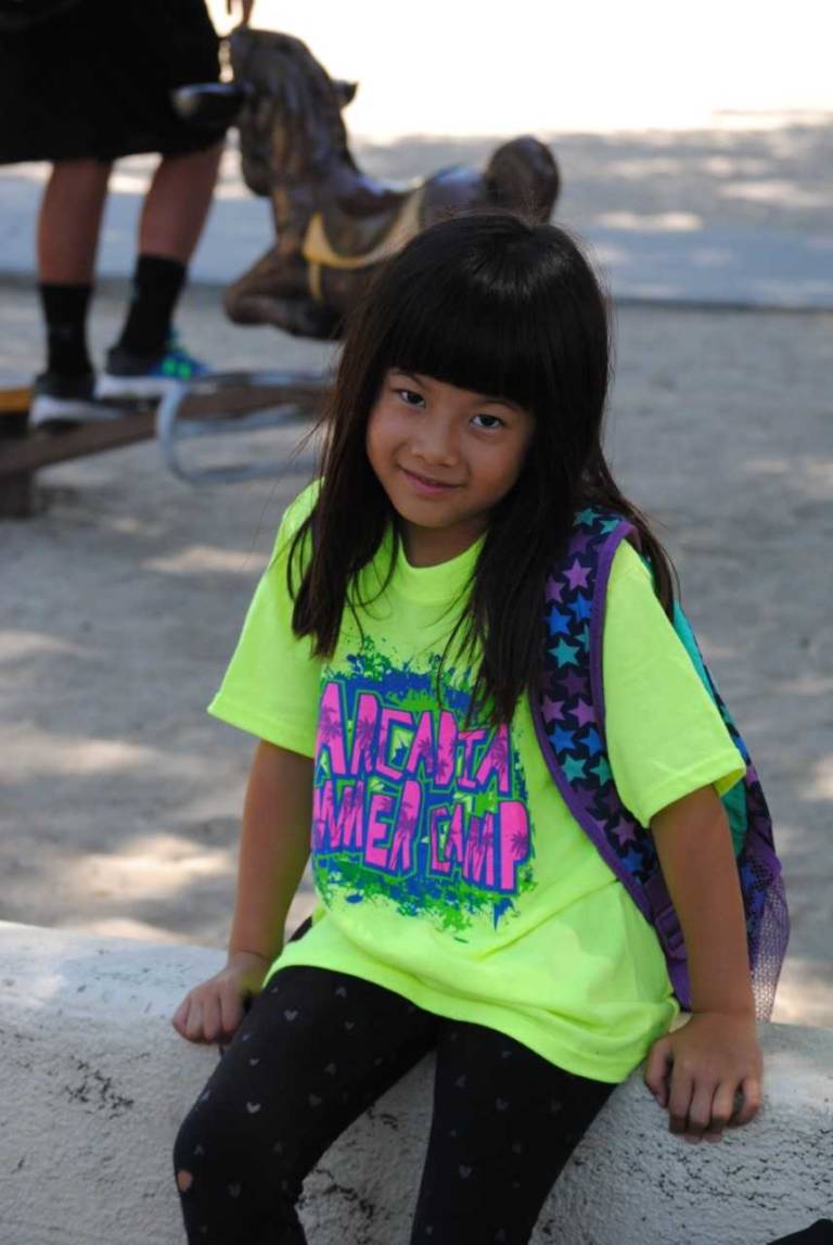 a young girl in a yellow shirt seated on a wall with a backpack on