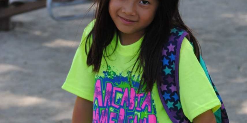 a young girl in a yellow shirt seated on a wall with a backpack on