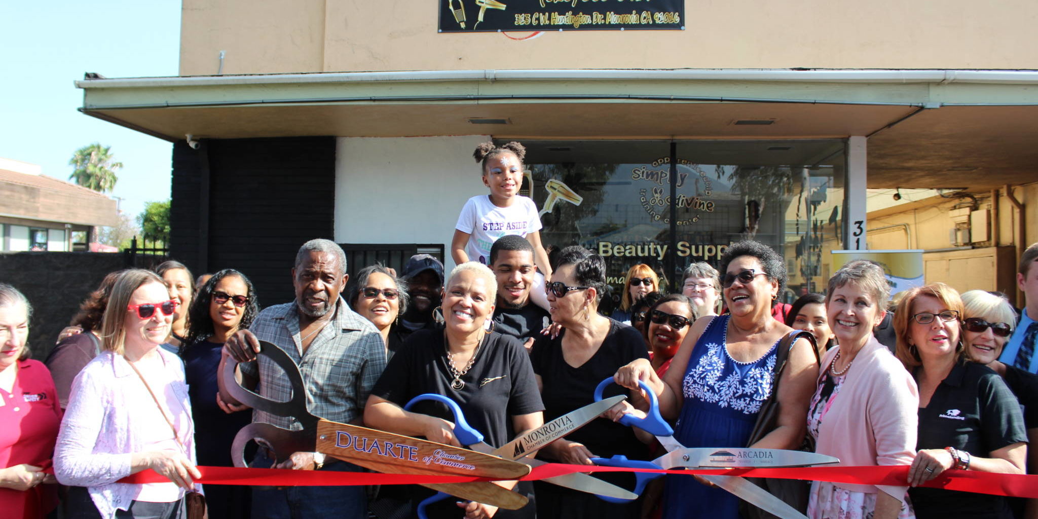 IMG_8689 a large group of people holding a pair of giant scissors and red ribbon beneath a sign for beauty supplies