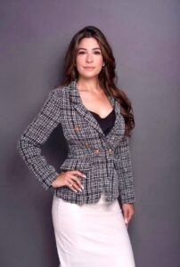 a woman in a white skirt wearing a plaid jacket over black top with long hair and one hand on a hip
