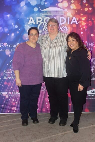 three women standing in front of a colorful step and repeat, and the taller woman in the center has her arms around the other two