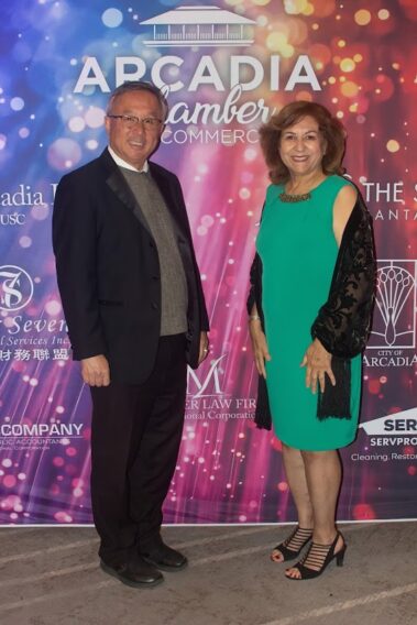 a woman in a green dress standing to the right of a man in a black suit, in front of a colorful step and repeat banner
