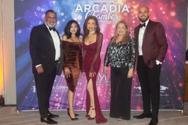 a group of three women flanked by two men, all standing in front of a colorful step and repeat banner