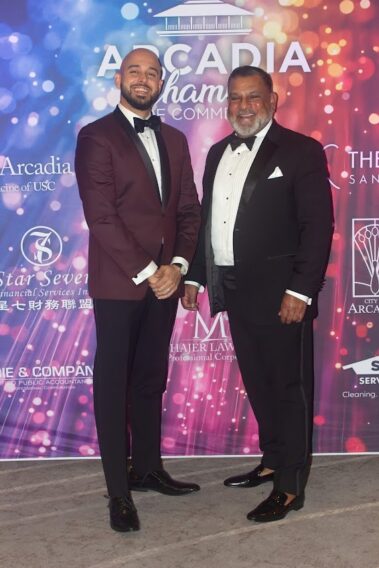 a man in a purple suit jacket standing next to a man in black suit, both in front of a colorful step and repeat banner