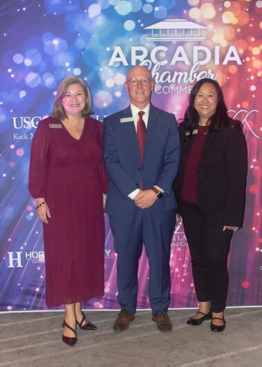 two women in business attire standing on either side of a man in a suit, all in front of a colorful step and repeat banner