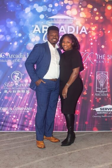 a woman in black attire standing beside a man in a blue suit with white shirt, in front of a colorful step and repeat banner