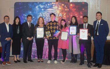 a man in a yellow and black checkered sweater standing in the middle of a group of people, holding plaques and certificates