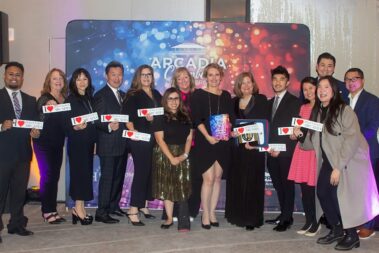a large group of people standing in front of a colorful step and repeat banner, all holding I love Downtown Arcadia stickers