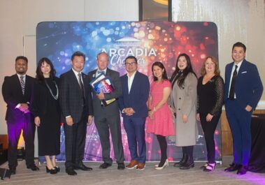 a man in a gray suit holding an armful of plaques, flanked by a group of women and men in front of a colorful step and repeat banner