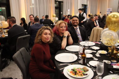 two women and a man seated at a table at an event with white plates in front of them on a black table cloth