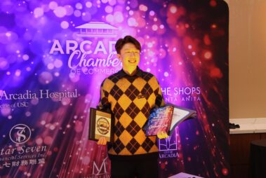 a man in a black and yellow checked sweated standing in front of a colorful step and repeat, holding awards