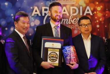 a tall man in black suit and colorful tie smiling while holding plaques and awards, standing between two shorter men