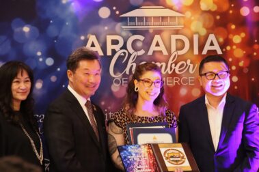 a woman in a leopard print dress holding certificates while standing between two men, in front of a colorful step and repeat