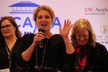 a woman in a black dress with her hair up holding a microphone with one hand up