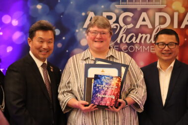 a woman holding a bunch of plaques and certificates while standing between two men, in front of a colorful step and repeat