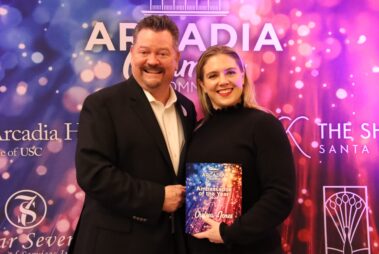 a woman and a man both wearing black standing in front of a colorful step and repeat, and she is holding a colorful plaque