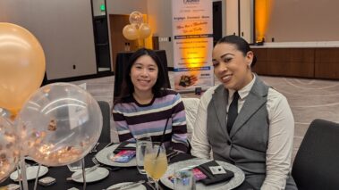 a woman in a purple striped sweater and a woman in a gray vest smiling at the camera while seated at a table at an event