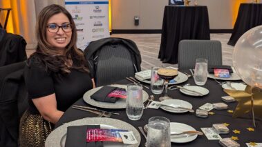 a woman in black seated at a table at an event set with white plates, black napkins, and colorful programs