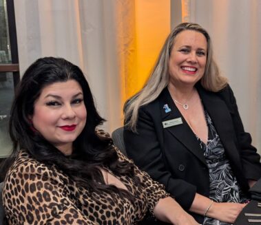 a woman with long black hair wearing a leopard print top seated with a woman with long, blonde hair in a black suit jacket