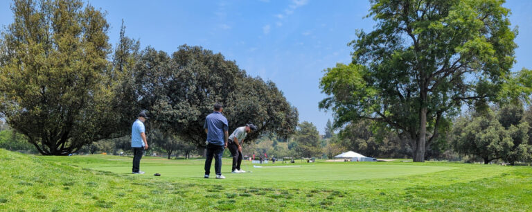 two male golfers watching another male golfer make a putt