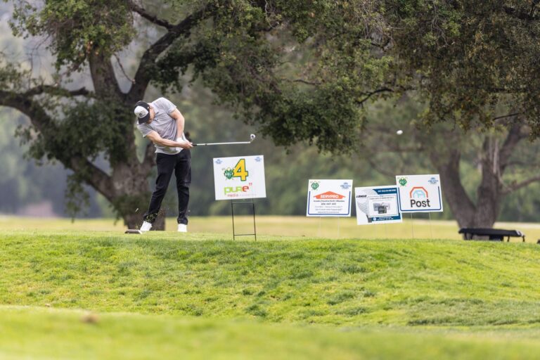 a man in a white shirt and black pants tees off in front of a line of tee sign s