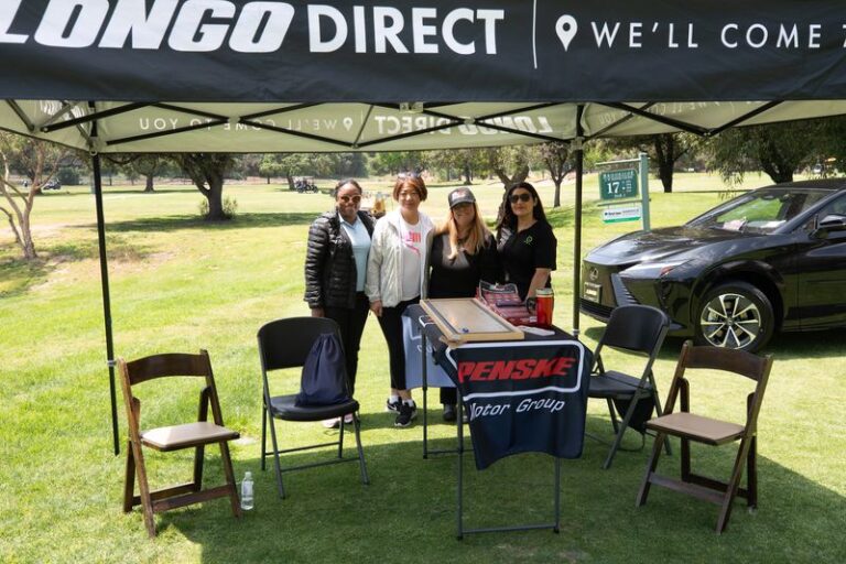 a group of women stand under a tent with the Longo Toyota logo on it and a black Lexus sedan behind them