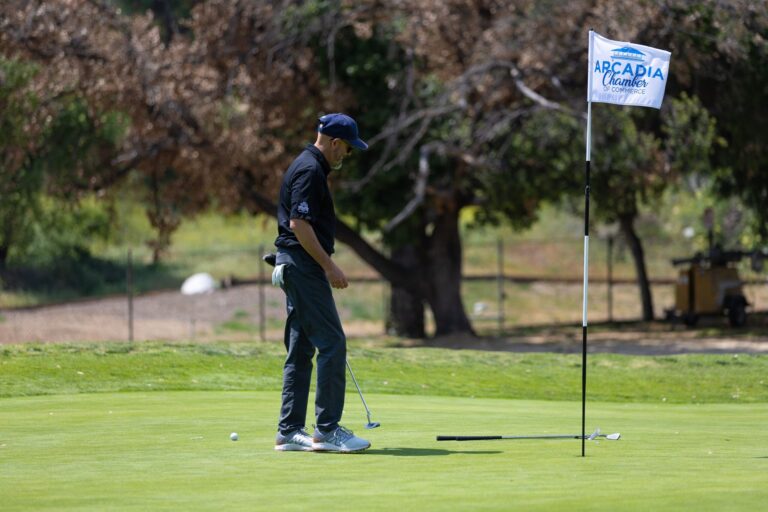 a man in black putts near a flag with the Arcadia chamber logo on it