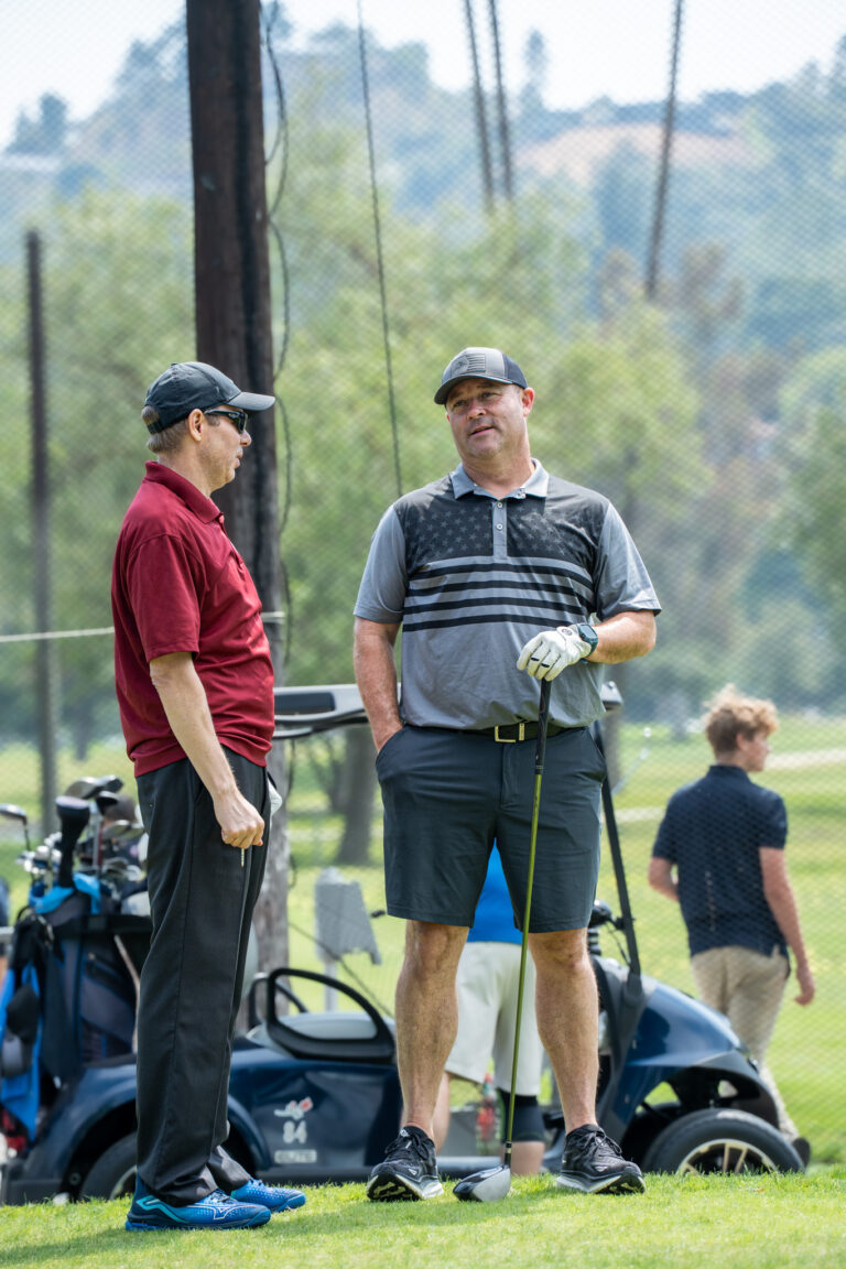 a man in red and a man in gray shirt talking to each other on a golf course