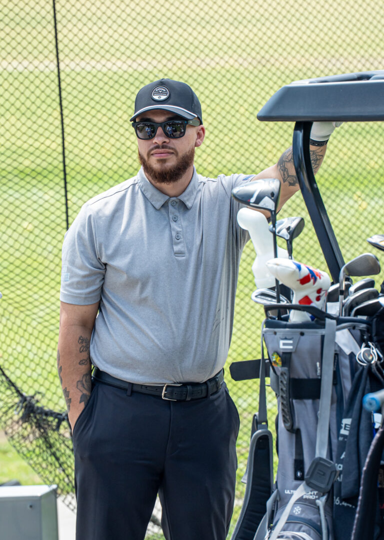a man in a gray shirt and black cap leaning on a golf cart full of golf clubs