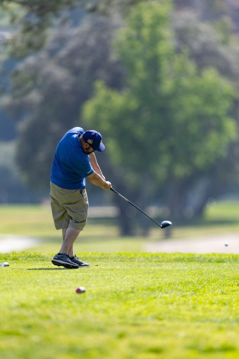 a man in a blue shirt and blue hat with khaki shorts tees off at a golf course