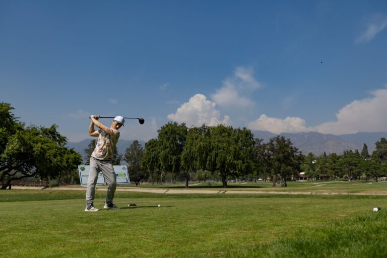 a man tees off at a golf course with the mountains and blue skies behind him