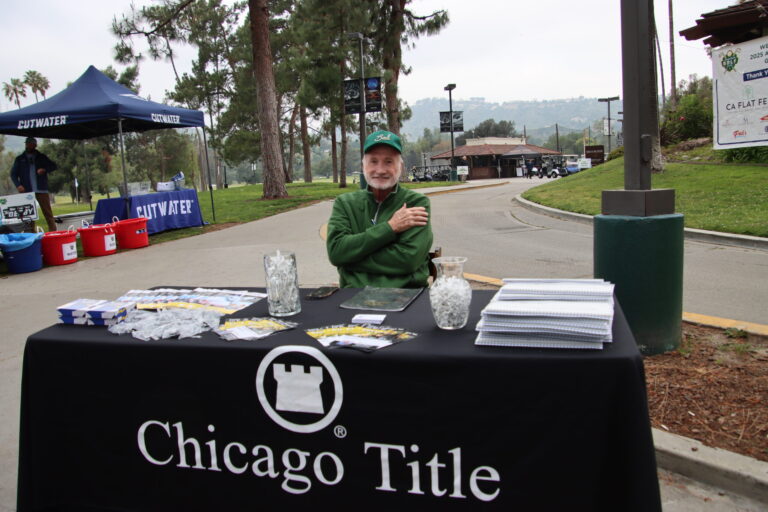 a table set with a black Chicago Title table cloth, with an older man seated behind it wearing a green jacket and green hat