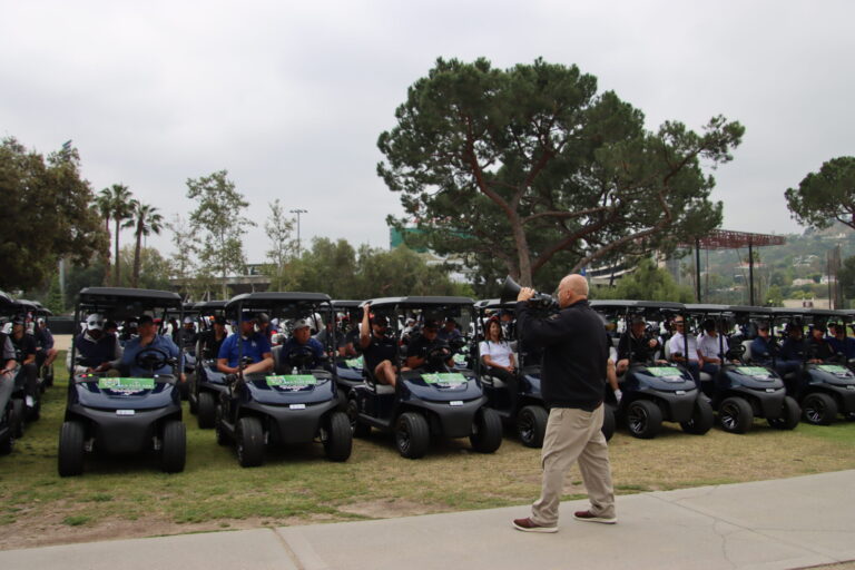 a man in a black shirt and khakis with a megaphone standing in front of a line of occupied golf carts