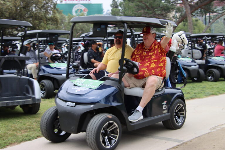 a man in a red patterned shirt in a golf cart waving, with a male companion in a yellow shirt in the other seat