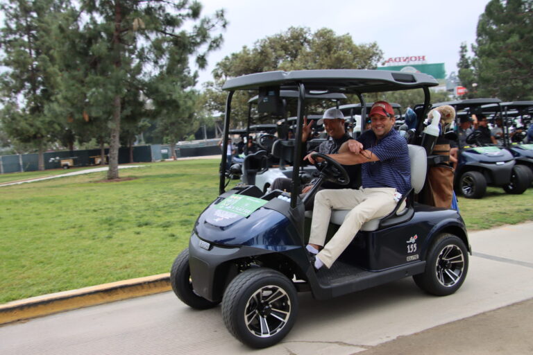 two men in a golf cart, with the driver pointing towards the camera