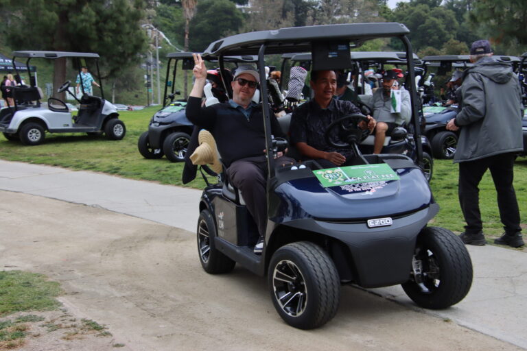 two men in a golf cart, with the passenger waving towards the camera
